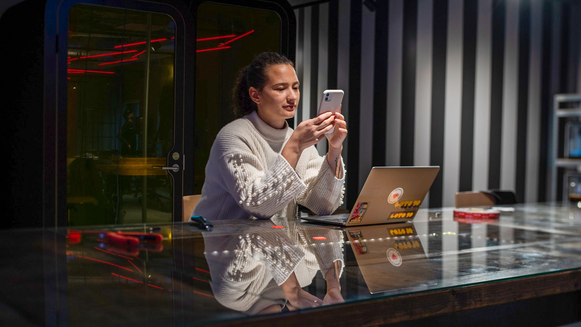 En kvinna sitter vid en dator med sin mobil på ett kontor.