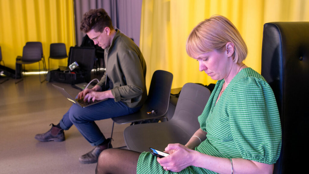 En kvinna i grön klänning sitter med sin mobil i handen, en man sitter med en laptop i knät.