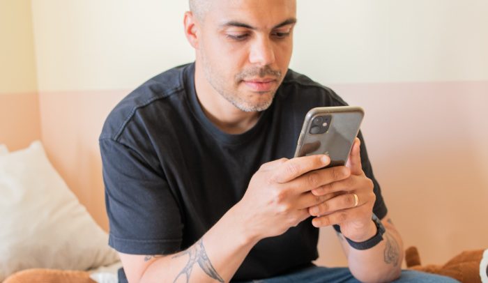 En man sitter på en säng och tittar på sin mobiltelefon.